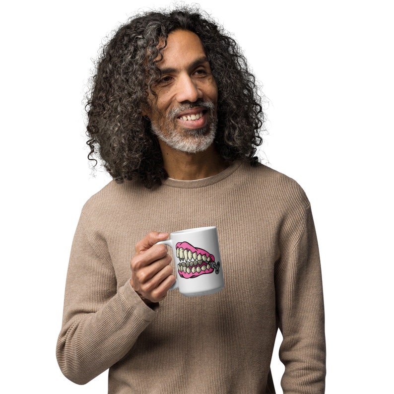 May include: A white coffee mug with a pink and yellow cartoon image of a set of dentures with a gold chain.