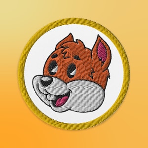 May include: A round, yellow embroidered patch with a cartoon fox face. The fox has orange fur, a white muzzle, and a pink tongue sticking out.