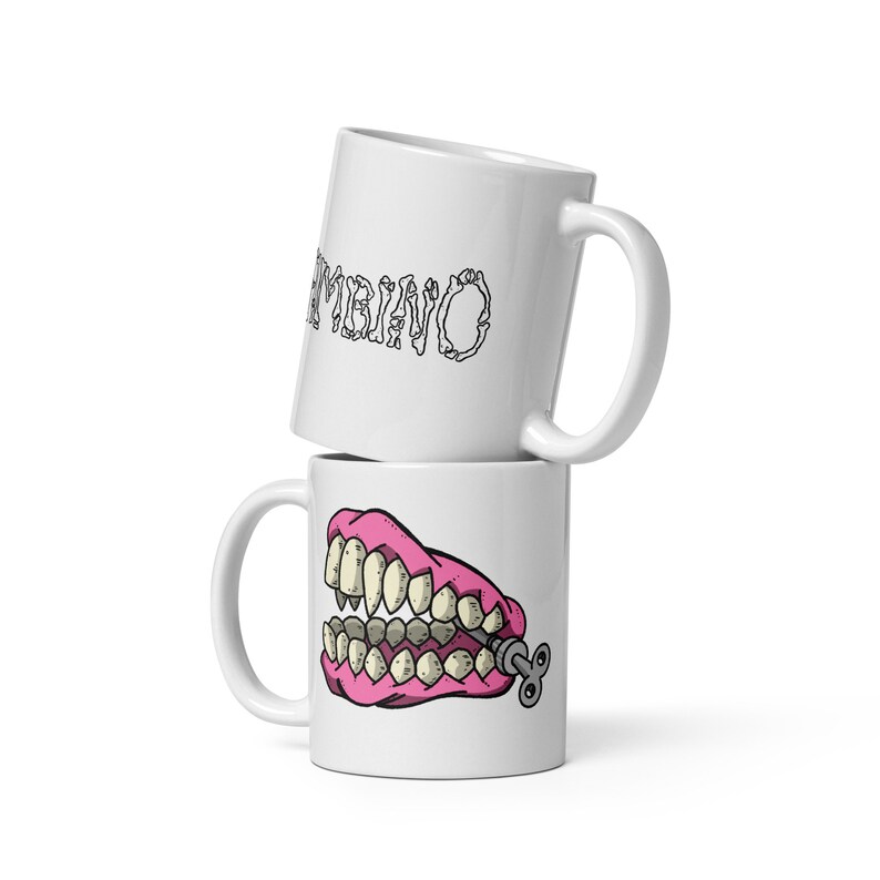 May include: Two white coffee mugs stacked on top of each other. The top mug has the word 'AMBIANO' written on it in a stylized font. The bottom mug has a cartoon image of a pink set of dentures with a winder key on the side.
