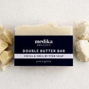 May include: A bar of soap with a navy blue label reading "DOUBLE BUTTER BAR" and "COCOA & SHEA BUTTER SOAP". The soap is flanked by pieces of cocoa butter and is branded "medika ORGANICS".