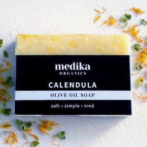 May include: A bar of Medika Organics Calendula Olive Oil Soap. The soap is light yellow with orange flecks. The black label has white text: "Calendula Olive Oil Soap" and "soft, simple, kind."