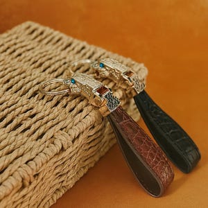 Può includere: Due portachiavi con un design a testa di leone color oro e un cinturino in pelle. Un portachiavi ha un cinturino marrone e l'altro un cinturino nero. Gli occhi del leone sono blu.