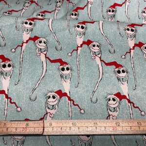 May include: Fabric featuring a repeating pattern of a tall, white character with a smiling face and a red Santa hat on a light blue background. The character has long, thin arms and legs. A wooden ruler is at the bottom.