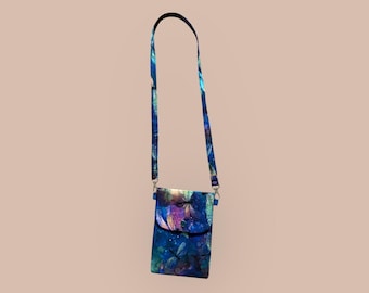 Puede incluir: Un bolso cruzado con cierre de solapa, con un vibrante estampado floral azul, morado y verde. La correa ajustable y el cuerpo del bolso muestran un diseño repetido de hojas y formas abstractas. El bolso está diseñado para llevar lo esencial.