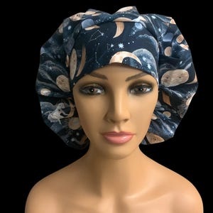 May include: A dark blue surgical scrub cap featuring a celestial design with moons, stars, and constellations. The fabric is patterned with various celestial bodies in shades of beige, white, and blue. The cap is designed to cover the head and hair.