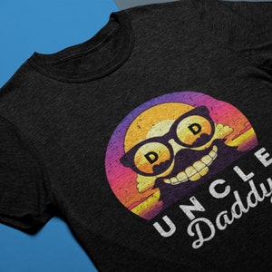 May include: A black t-shirt featuring a retro-style graphic of a smiling face wearing sunglasses with the letters "D" on the lenses. The graphic is set against a sunset-coloured background. The text "UNCLE Daddy" is printed below the graphic in white.
