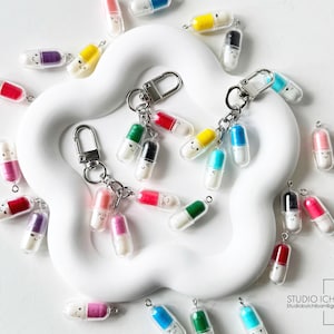 May include: A collection of colorful pill-shaped keychains with smiling faces. The keychains feature various colors, including red, yellow, green, and blue, and are attached to silver key rings. The keychains are scattered on a white, flower-shaped surface.