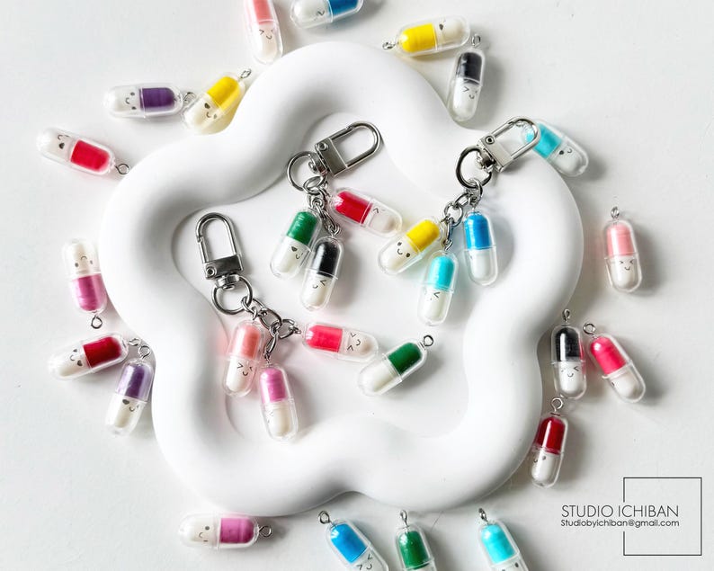 May include: A collection of colorful pill-shaped keychains with smiling faces. The keychains feature various colors, including red, yellow, green, and blue, and are attached to silver key rings. The keychains are scattered on a white, flower-shaped surface.