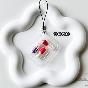 May include: A clear plastic blister pack containing three colorful capsule-shaped pills. The pack is attached to a black string and sits on a white, wavy, flower-shaped tray. The text "TO GO PILLS" is visible.