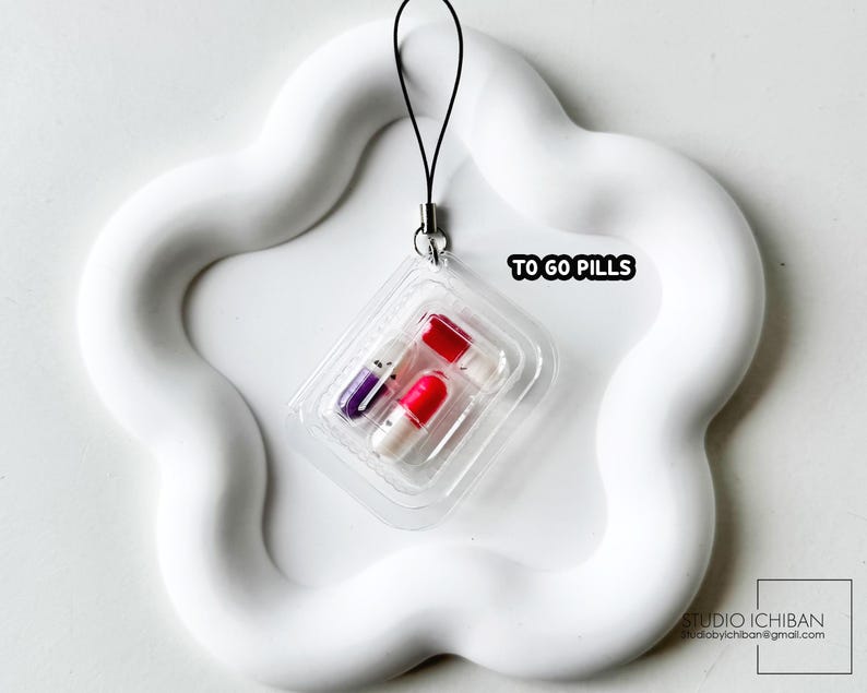 May include: A clear plastic blister pack containing three colorful capsule-shaped pills. The pack is attached to a black string and sits on a white, wavy, flower-shaped tray. The text "TO GO PILLS" is visible.
