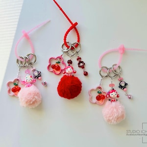 May include: Three pink and red keychains with pom-poms, charms, and key rings. Each keychain features a small doll with a cherry and a flower charm. The keychains are attached to pink and red cords.