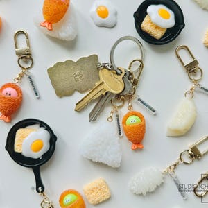 Puede incluir: Un conjunto de llaveros con comida en miniatura, incluyendo huevos fritos, bolas de arroz y pollo frito. Los llaveros est&aacute;n hechos de resina colorida y tienen un dise&ntilde;o juguet&oacute;n y caprichoso. Los llaveros est&aacute;n unidos a anillos y cadenas de metal dorado.