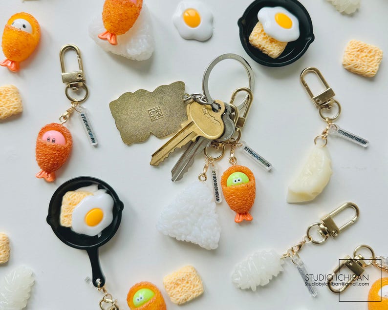 Puede incluir: Un conjunto de llaveros con comida en miniatura, incluyendo huevos fritos, bolas de arroz y pollo frito. Los llaveros est&aacute;n hechos de resina colorida y tienen un dise&ntilde;o juguet&oacute;n y caprichoso. Los llaveros est&aacute;n unidos a anillos y cadenas de metal dorado.