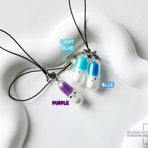 May include: Three colorful pill-shaped charms with smiling faces, each attached to a black lanyard. The charms are light blue, blue, and purple, with the words "LIGHT BLUE", "BLUE", and "PURPLE" next to each charm. The background is a white, wavy surface.