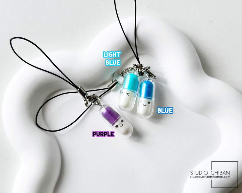 May include: Three colorful pill-shaped charms with smiling faces, each attached to a black lanyard. The charms are light blue, blue, and purple, with the words "LIGHT BLUE", "BLUE", and "PURPLE" next to each charm. The background is a white, wavy surface.
