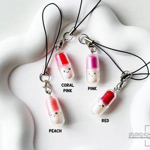 May include: Four colorful pill-shaped charms with smiling faces, each attached to a black lanyard. The charms are in coral pink, pink, peach, and red. The text on the image reads "CORAL PINK", "PINK", "PEACH", and "RED". The Studio Ichiban logo is in the bottom right corner.