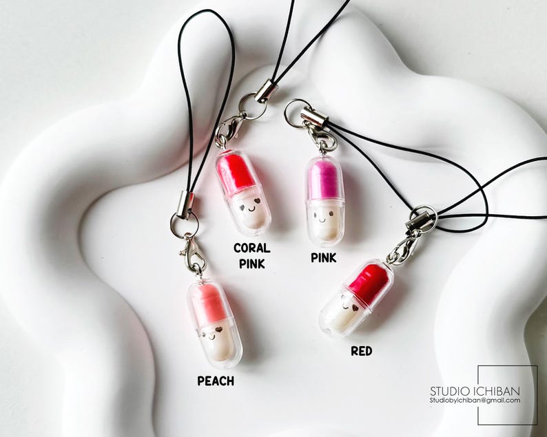 May include: Four colorful pill-shaped charms with smiling faces, each attached to a black lanyard. The charms are in coral pink, pink, peach, and red. The text on the image reads "CORAL PINK", "PINK", "PEACH", and "RED". The Studio Ichiban logo is in the bottom right corner.