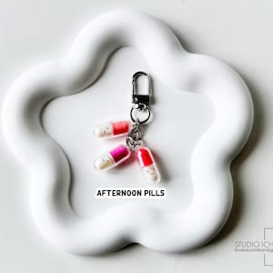 May include: A keychain featuring three colorful pill charms with smiling faces. The pills are red, pink, and orange, attached to a silver clasp. The text "AFTERNOON PILLS" is printed below the charms.