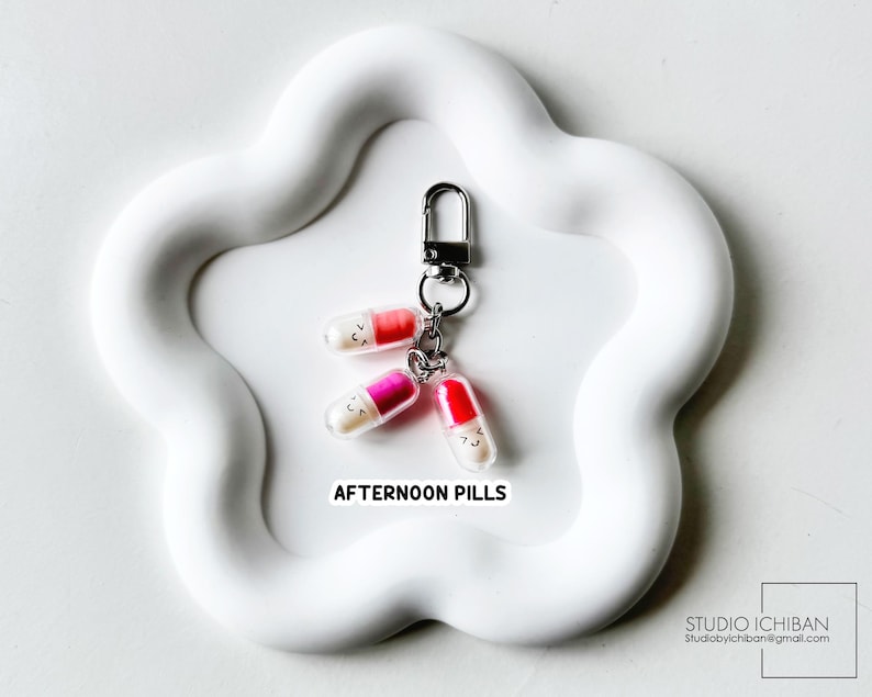 May include: A keychain featuring three colorful pill charms with smiling faces. The pills are red, pink, and orange, attached to a silver clasp. The text "AFTERNOON PILLS" is printed below the charms.