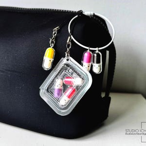 May include: A black cosmetic bag with a white ring and several capsule-shaped charms. The charms are in various colors, including yellow, pink, and purple, and have smiling faces. A clear plastic container holds additional capsule charms.