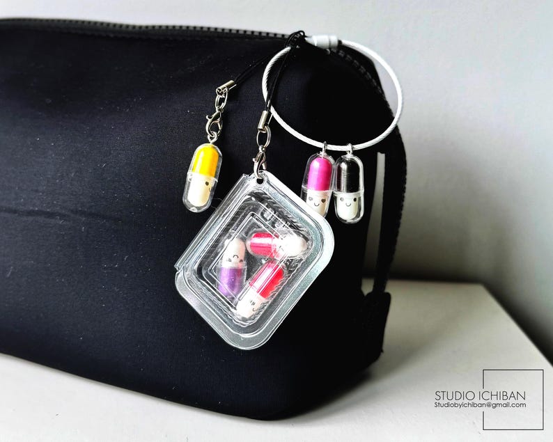 May include: A black cosmetic bag with a white ring and several capsule-shaped charms. The charms are in various colors, including yellow, pink, and purple, and have smiling faces. A clear plastic container holds additional capsule charms.