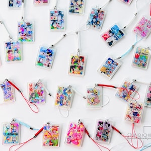 May include: A collection of rectangular acrylic charms with colorful anime-style illustrations. Each charm is attached to a lanyard with a white or colored cord. The charms feature various characters and scenes, with a white background.