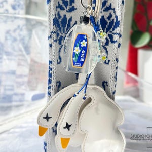 May include: A clear, rectangular keychain with a label featuring white flowers and Japanese text. The keychain is attached to a white leather goose foot charm with yellow beaks and black crosses. A blue and white patterned fabric is in the background.
