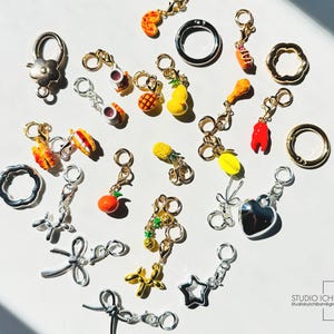 May include: A collection of assorted metal charms featuring miniature food items like fruit, fried chicken, and lobster, alongside shapes including stars, hearts, bows, and balloon animals, all equipped with small lobster claw clasps for attachment.