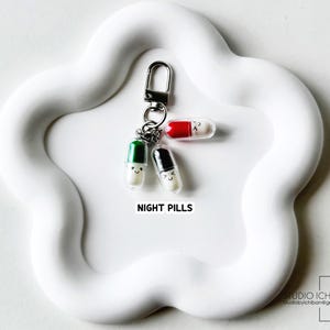 May include: A keychain featuring three smiling pill charms with different colored caps: green, black, and red. The charms are attached to a silver clasp and are on a white, cloud-shaped tray. The text "NIGHT PILLS" is below the charms.