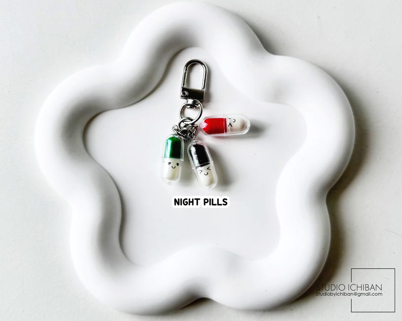 May include: A keychain featuring three smiling pill charms with different colored caps: green, black, and red. The charms are attached to a silver clasp and are on a white, cloud-shaped tray. The text "NIGHT PILLS" is below the charms.