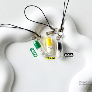 May include: Three colorful pill-shaped charms with smiling faces, each attached to a black lanyard. The charms are green, yellow, and black, with the colors labeled. The background is a white, wavy surface.