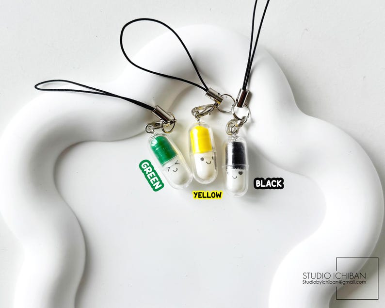 May include: Three colorful pill-shaped charms with smiling faces, each attached to a black lanyard. The charms are green, yellow, and black, with the colors labeled. The background is a white, wavy surface.