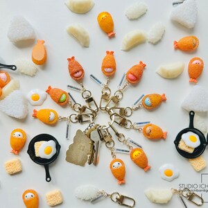 Puede incluir: Una colecci&oacute;n de llaveros coloridos que representan diferentes alimentos japoneses, como huevos fritos, tempura de camarones y sushi. Los llaveros est&aacute;n hechos de arcilla polim&eacute;rica y tienen un estilo lindo y de dibujos animados. Los llaveros se muestran sobre un fondo blanco.