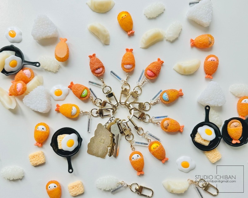 Puede incluir: Una colecci&oacute;n de llaveros coloridos que representan diferentes alimentos japoneses, como huevos fritos, tempura de camarones y sushi. Los llaveros est&aacute;n hechos de arcilla polim&eacute;rica y tienen un estilo lindo y de dibujos animados. Los llaveros se muestran sobre un fondo blanco.
