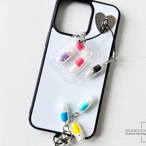 May include: A white iPhone case with a black border, featuring a heart-shaped attachment and a charm with pill-shaped pendants in various colors. The charm includes a clear plastic blister pack with colorful pills.