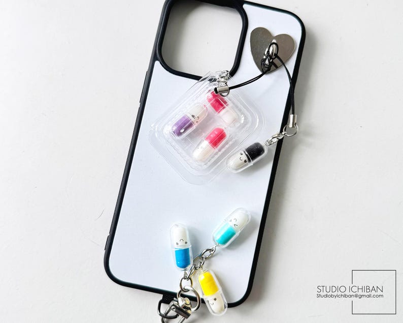 May include: A white iPhone case with a black border, featuring a heart-shaped attachment and a charm with pill-shaped pendants in various colors. The charm includes a clear plastic blister pack with colorful pills.