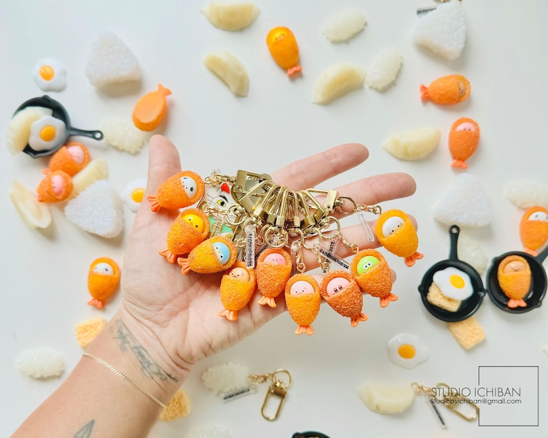 Puede incluir: Una colecci&oacute;n de abalorios de llavero naranja y amarillo con forma de alimentos japoneses, incluyendo un huevo frito, una bola de arroz y un rollo de sushi. Los abalorios est&aacute;n unidos a llaveros de oro y se muestran en una mano.
