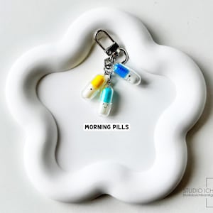 May include: A keychain with three colorful pill charms, each with a smiling face. The pills are yellow, blue, and turquoise, attached to a silver clasp. The text "MORNING PILLS" is visible on a white, flower-shaped surface.