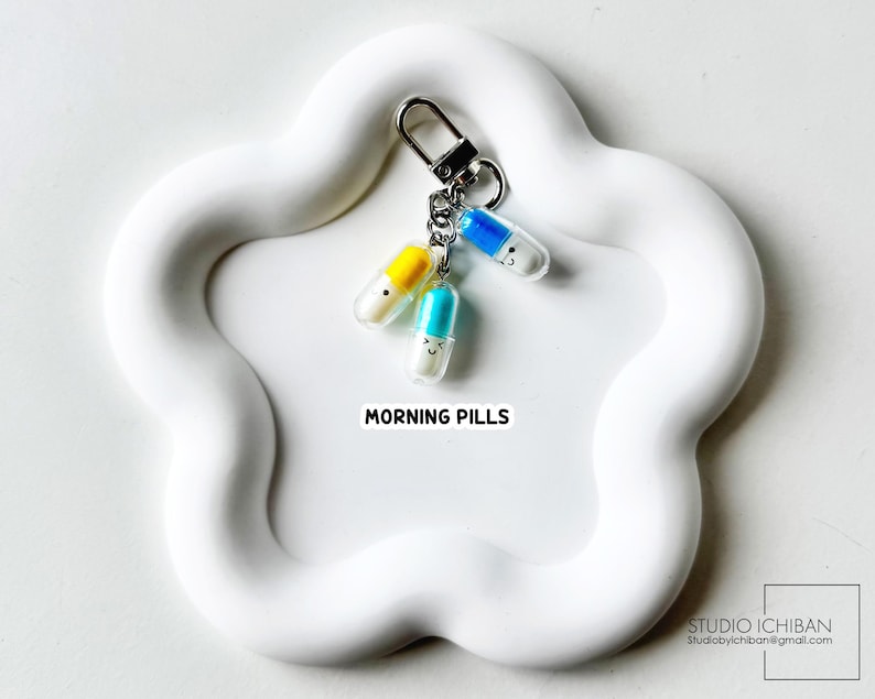 May include: A keychain with three colorful pill charms, each with a smiling face. The pills are yellow, blue, and turquoise, attached to a silver clasp. The text "MORNING PILLS" is visible on a white, flower-shaped surface.