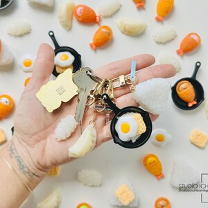 Puede incluir: Un llavero con una variedad de amuletos de comida en miniatura, incluyendo huevos fritos, bolas de arroz y nuggets de pollo. Los amuletos est&aacute;n unidos a un llavero dorado y un amuleto de sart&eacute;n negra.