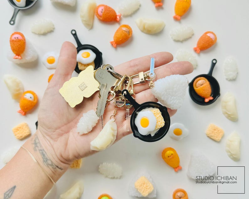 Puede incluir: Un llavero con una variedad de amuletos de comida en miniatura, incluyendo huevos fritos, bolas de arroz y nuggets de pollo. Los amuletos est&aacute;n unidos a un llavero dorado y un amuleto de sart&eacute;n negra.