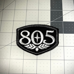 May include: A black and white embroidered patch with the number 805 and a star in the center. The patch is shaped like a shield and has a white embroidered border.