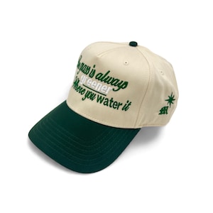 May include: Cream-colored baseball cap with a dark green brim and top button. The cap features the embroidered phrase "The grass is always greener where you water it" in green script. A small green star and logo are on the side.