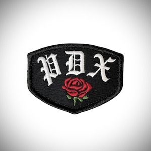 May include: Black and white embroidered patch with the text "PDX" in a gothic font. A red rose with green leaves is centered below the text.