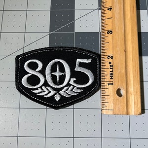 May include: Black and white embroidered patch with the number 805 and a star in the center. The patch is shaped like a shield and has a white embroidered design around the number.