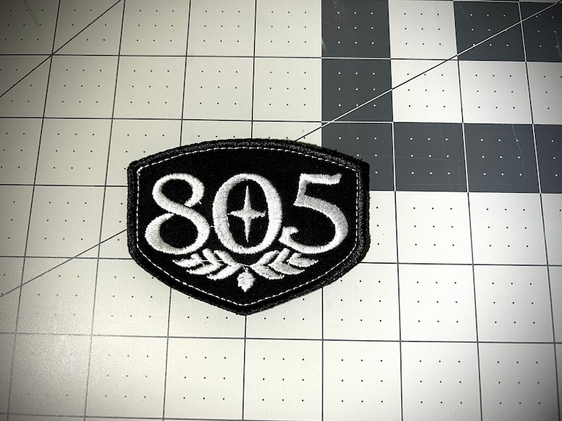 May include: A black and white embroidered patch with the number 805 and a star in the center. The patch is shaped like a shield and has a white embroidered border.