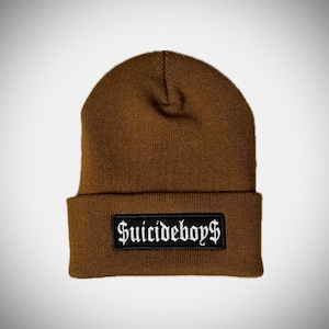 May include: A brown knit beanie with a folded brim. A black rectangular patch with the text "SUICIDEBOYS" in white gothic lettering is sewn onto the folded brim of the hat.