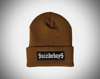 May include: A brown knitted beanie hat with a turned-up brim. The folded brim features a black rectangular patch with the white gothic text "SUICIDEBOYS".