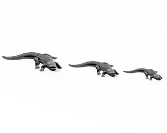 May include: Three gray alligator figurines of varying sizes are arranged in a row. The alligators are depicted in a walking pose, with detailed scales and open mouths. The background is plain white.