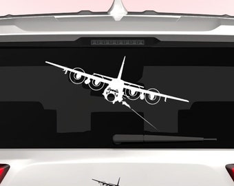 Könnte beinhalten: Ein weißes Auto mit einer weißen Silhouette eines Flugzeugs auf der Heckscheibe. Das Flugzeug fliegt mit einem Seil, das hinter ihm herhängt. Das Auto hat einen "DINGO"-Aufkleber auf der hinteren Stoßstange und einen dünnen blau-weißen amerikanischen Flaggenaufkleber auf der rechten unteren Seite der Stoßstange.